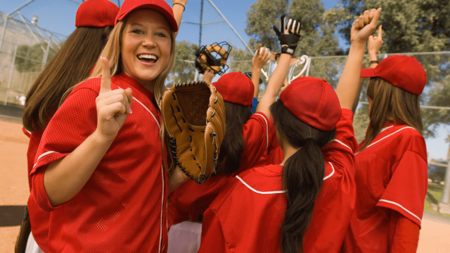 A softball player facing the camera and holding a finger up celebrating with her team
